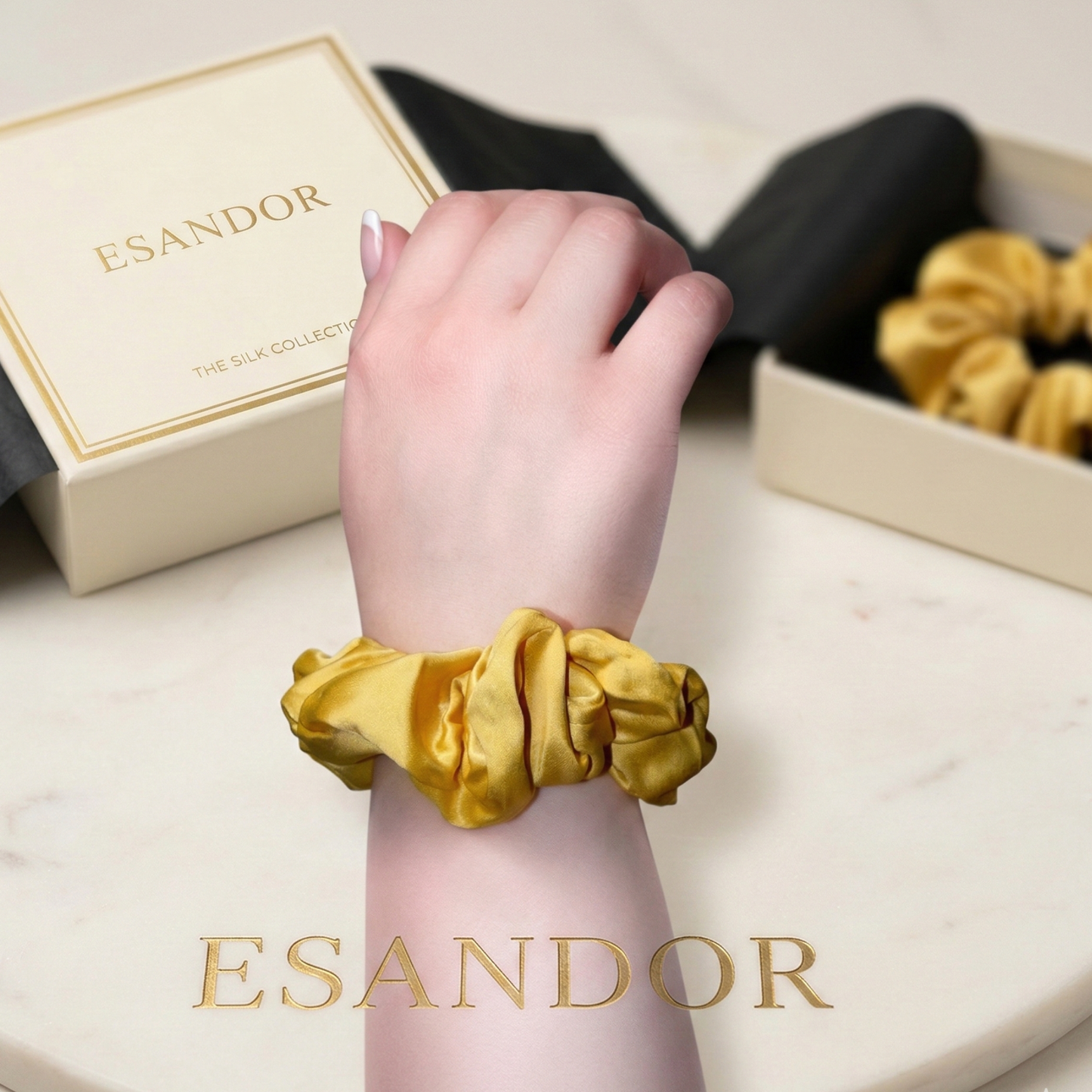 Gold scrunchie on a wrist with 'ESANDOR' branding in the background