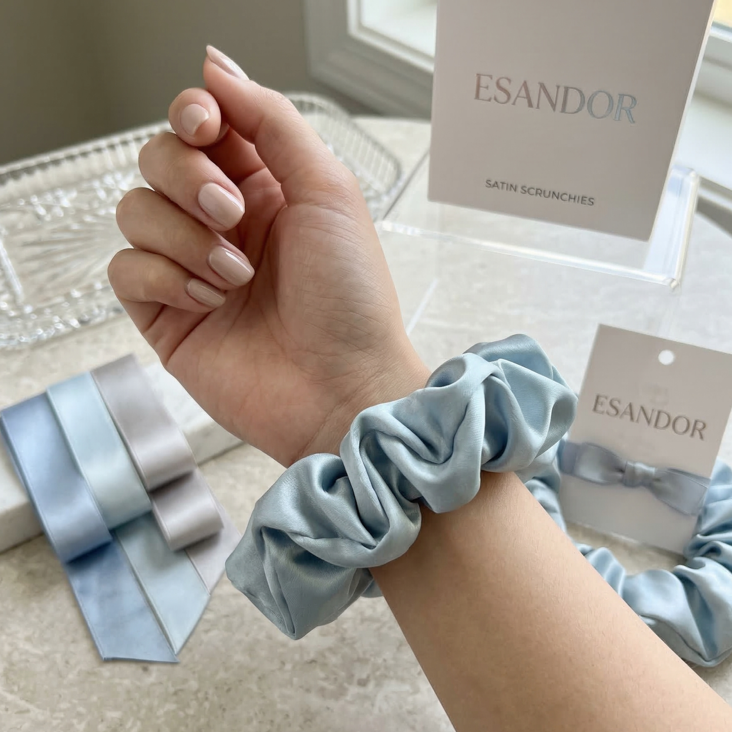Hand wearing a light blue satin scrunchie with 'ESANDOR' branding in the background.