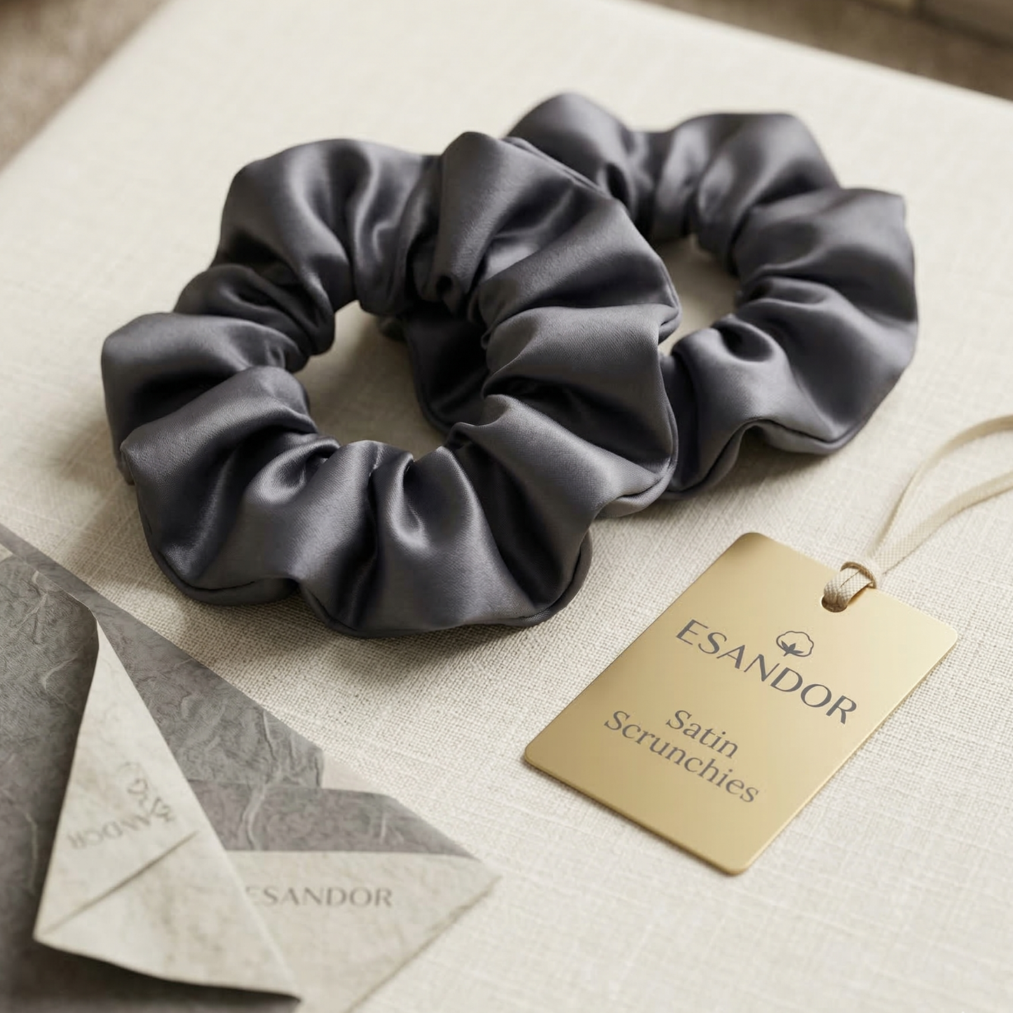 Two gray satin scrunchies with an Esandor tag on a light fabric background
