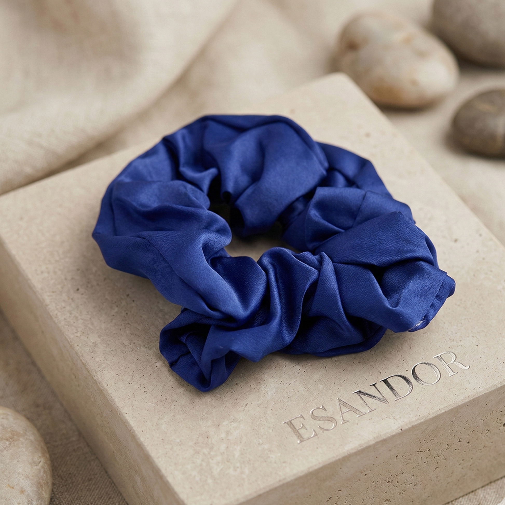 Blue scrunchie on a beige block with 'ESANDOR' branding, surrounded by stones.