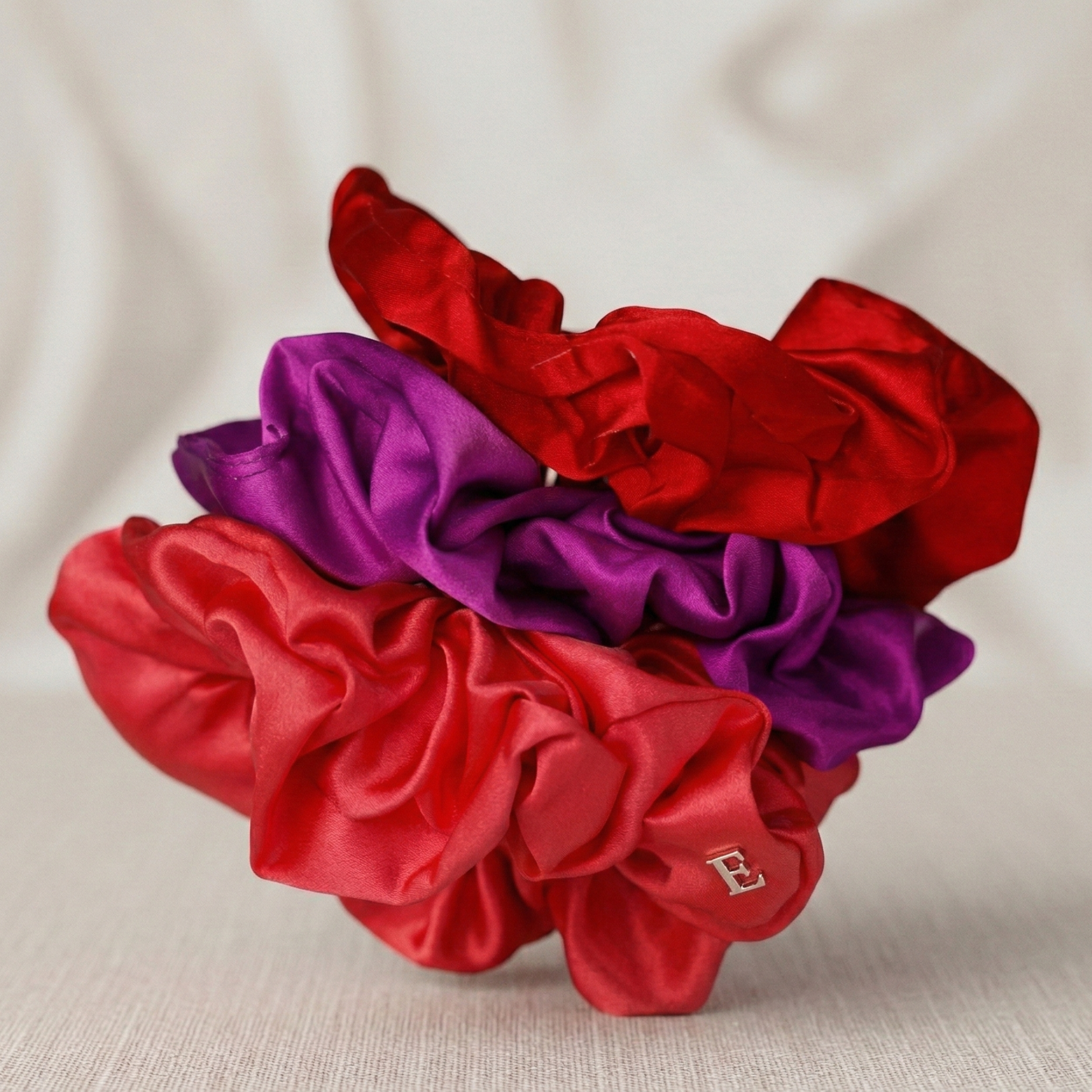Three satin scrunchies in red and purple on a beige fabric background